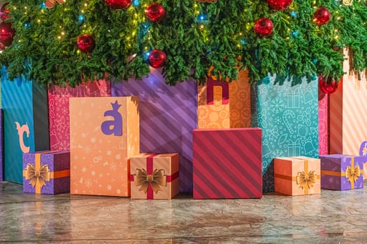 Brightly wrapped gifts in various sizes sit under a beautifully decorated Christmas tree. The vibrant colors and patterns create a cheerful holiday atmosphere.