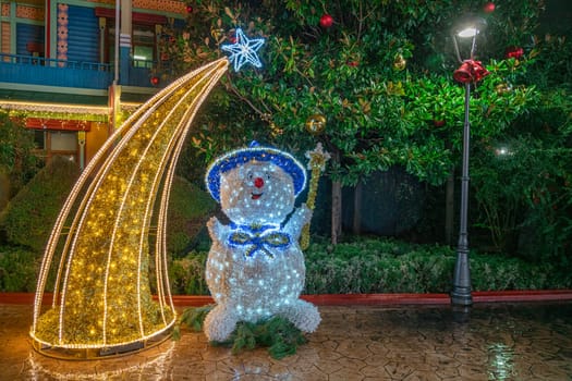 A cheerful, illuminated snowman stands next to a sparkling star and tree in the park, adorned with colorful lights and surrounded by lush greenery during the holiday season.