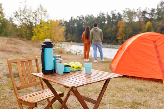 A camping scene with a wooden table set with a thermos, cups, and apples. An orange tent is pitched nearby, and a couple stands by a lake, surrounded by autumn trees.