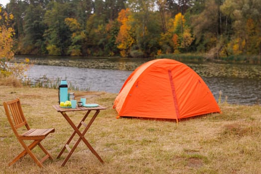 A vibrant orange tent set up by a serene riverbank, surrounded by autumn foliage. A wooden table with a thermos, cups, and apples is nearby, creating a cozy camping scene.