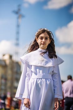 Young girl is standing outdoors in a white dress holding a rosary for her first communion ceremony
