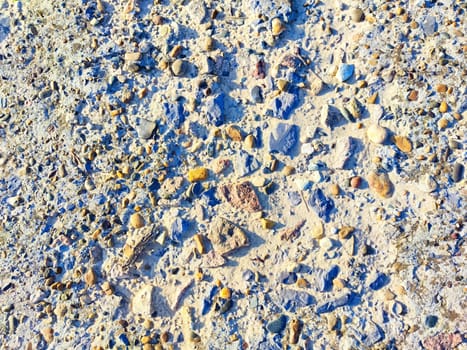 A variety of smooth stones in different colors are scattered on a sandy ground, illuminated by sunlight in an outdoor environment.