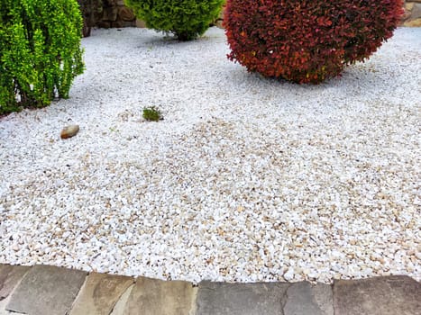 A serene garden showcases white gravel ground surrounded by lush, well-manicured shrubs in various hues.