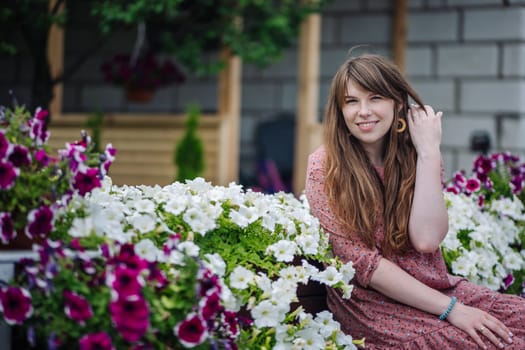 Young woman with long brown hair sits on a bench surrounded by white and purple flowers, adjusting her hair with a smile