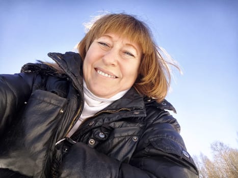 A cheerful middle aged woman in a winter coat taking selfie on nature outdoors in sunny day with blue sky