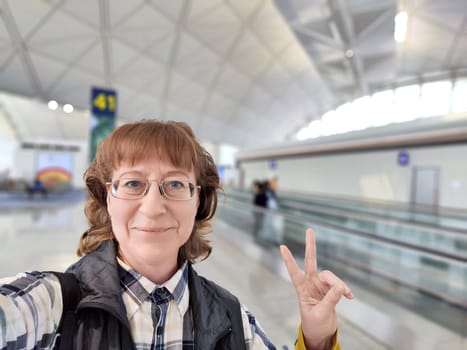 A cheerful middle-aged woman poses for a selfie at an airport lounge, eagerly awaiting her next flight departure.