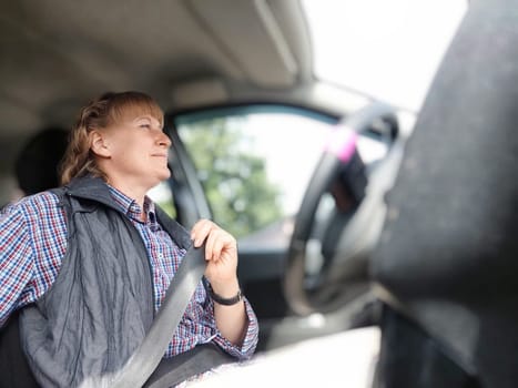 Middle aged woman is seated in a car, smiling cheerfully as she securely fastens her seat belt. Safety in transport. Traffic Laws