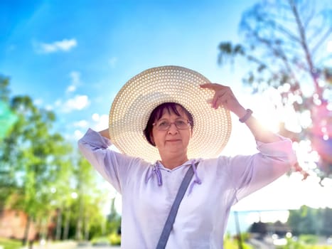 A woman is playfully adjusting her large sun hat while standing in vibrant park filled with trees under a clear blue sky