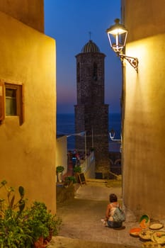 Woman sitting on steps looking at a beautiful view of Castelsardo at dusk. Sardinia, Italy
