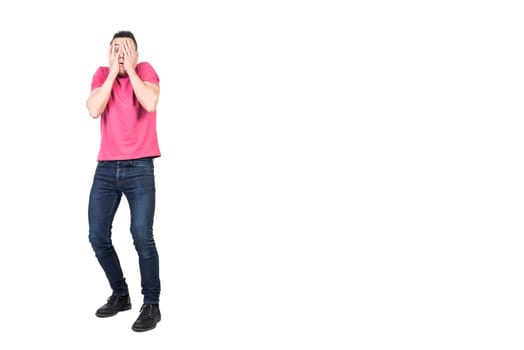 Full length scared man in casual clothes covering face with hands and peeking through fingers isolated on white background