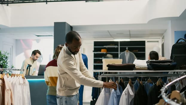 Young adult examining materials of formal wear hanging in clothing store at shopping center. Male client looking at trendy modern clothes or merchandise, buying fashion brands in retail shop.