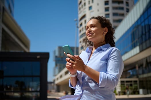 Beautiful female manager in casual wear, entrepreneur, freelancer, businesswoman using mobile phone, smiling and looking confidently aside, standing against modern office urban buildings background