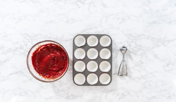 Flat lay. Scooping cupcake dough with dough scoop into cupcake pan lined with foil liners to bake red velvet cupcakes with white chocolate ganache frosting.