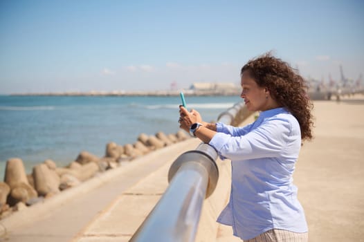 Positive African American woman holding mobile phone, relaxing standing at seacoast on a beautiful summer sunny day, dressed in blue casual shirt, browsing websites, checking social media content