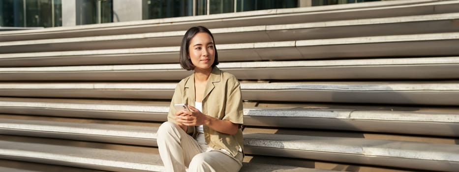 Beautiful asian girl sitting on stairs outside building, using mobile phone, looking at smartphone app and smiling.