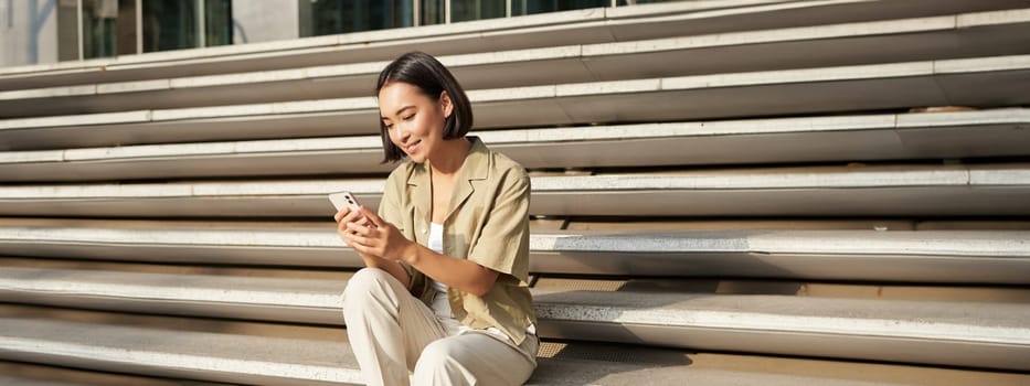 Beautiful asian girl sitting on stairs outside building, using mobile phone, looking at smartphone app and smiling.