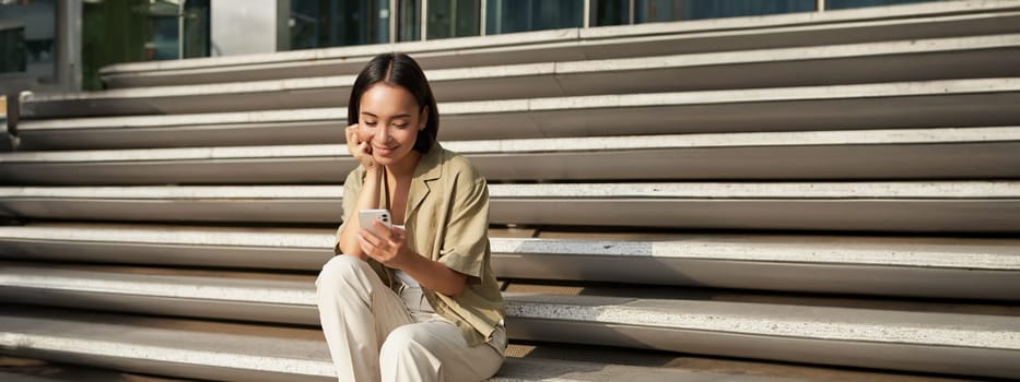 Vertical shot of asian woman, student sits on stairs in city, looking at mobile phone screen and smiling, using smartphone app.