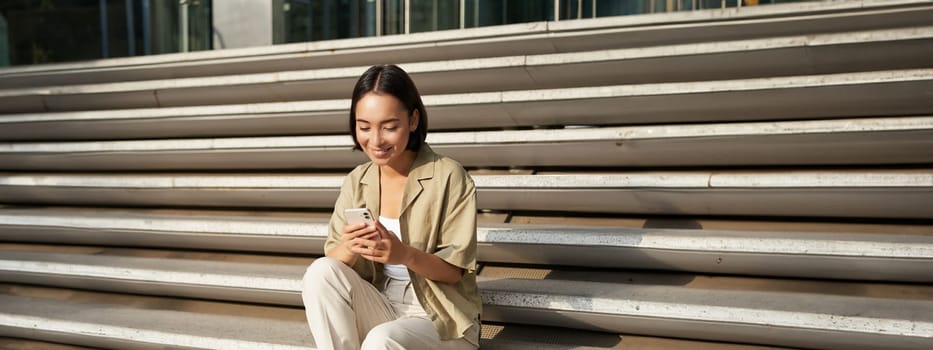 Beautiful asian girl sitting on stairs outside building, using mobile phone, looking at smartphone app and smiling.