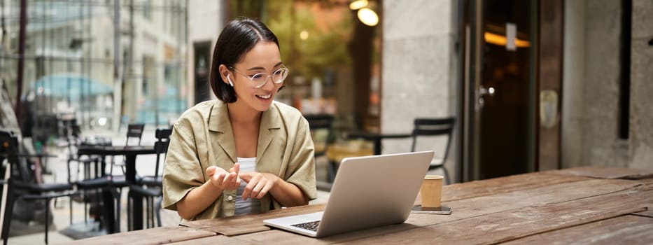 Young woman sitting on online meeting in outdoor cafe, talking to laptop camera, explaining something, drinking coffee. Digital nomad