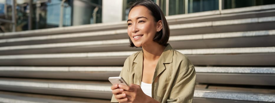 People and technology. Smiling beautiful asian woman sitting on stairs in city, holding mobile phone. Girl with smartphone rests outside.