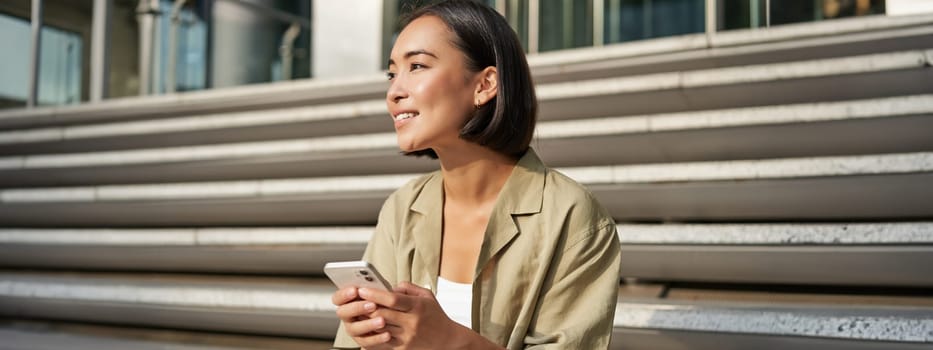 People and technology. Smiling beautiful asian woman sitting on stairs in city, holding mobile phone. Girl with smartphone rests outside.