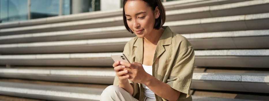 People and technology. Smiling beautiful asian woman sitting on stairs in city, holding mobile phone. Girl with smartphone rests outside.