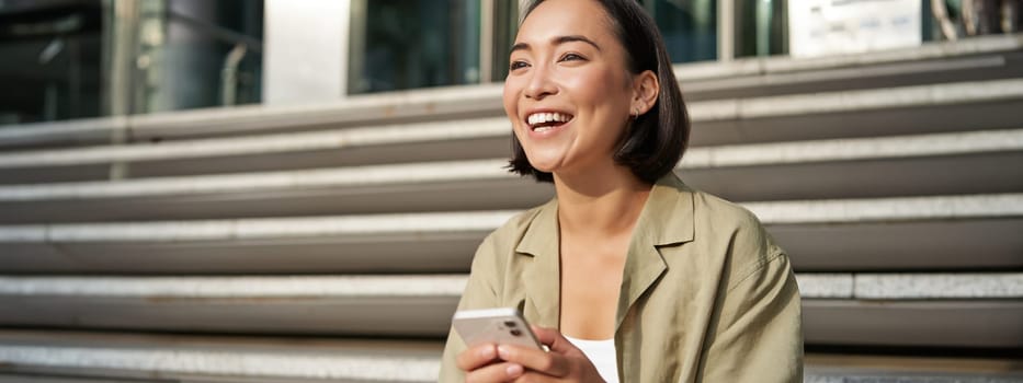 Beautiful asian girl sits on stairs with smartphone. Young korean woman resting outdoors, using mobile phone application.