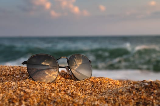 On the beach, sunglasses lie on the sand in the background a wave rolls from the sea. Close-up of leisure accessory, no people.