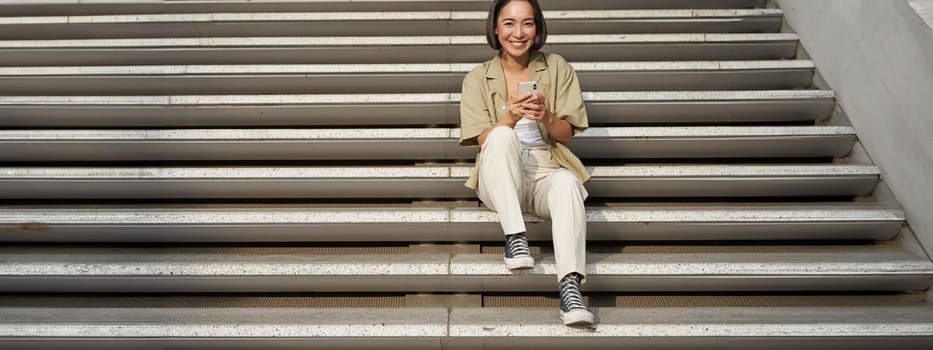 Portrait of smiling asian girl sits on stairs with her smartphone, browsing internet on mobile phone, resting outdoors in city centre.