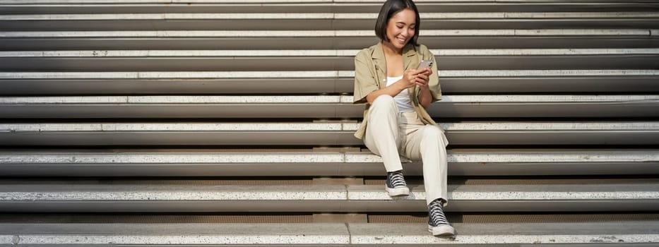 Portrait of smiling asian girl sits on stairs with her smartphone, browsing internet on mobile phone, resting outdoors in city centre.