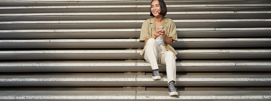 Portrait of smiling asian girl sits on stairs with her smartphone, browsing internet on mobile phone, resting outdoors in city centre.