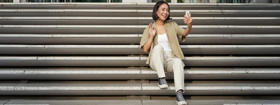 Portrait of asian beautiful girl takes selfie on smartphone. Young korean woman sits on stairs outdoors and makes photos on mobile phone.