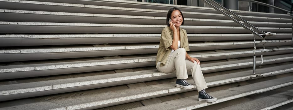 Smiling asian girl sits on stairs of building and talks on mobile phone, relaxing during telephone conversation.