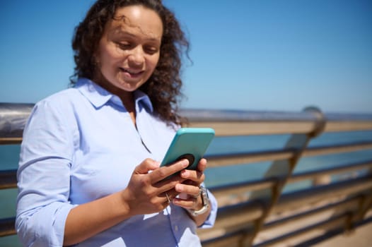 Close-up of a female manager, entrepreneur holding her modern smartphone, checking new mobile app, booking, ordering, scrolling newsfeed, typing text on digital screen, against Ocean background