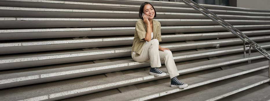 Portrait of asian student girl on stairs talks on mobile phone, smiles at camera, sits outside building.