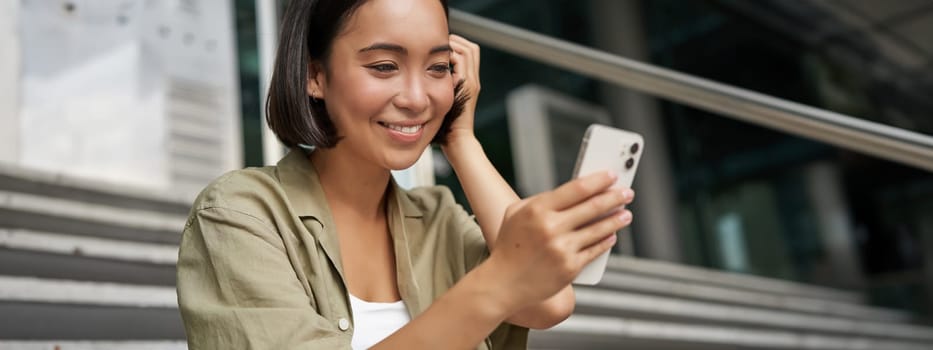 Portrait of asian girl takes selfie on mobile phone. Korean woman smiling, video chat on smartphone app while sits outside on stairs.