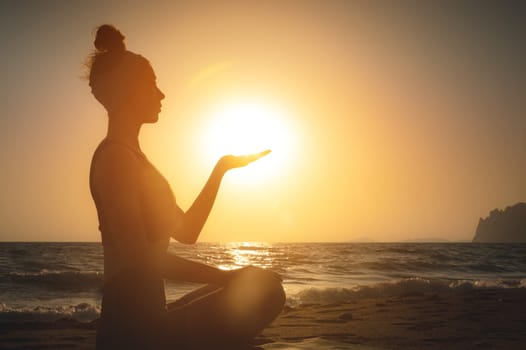 Side view. Silhouette shot at sunset or sunrise. Young caucasian woman meditating in the lotus position on the seashore holds the sun on her hand. Yoga and meditation practice at sunrise by the sea.