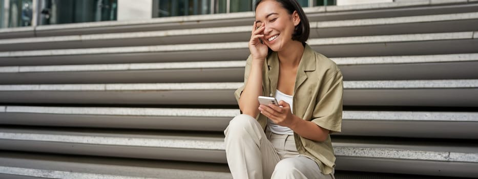 Portrait of happy asian girl sits with mobile phone and laughs. Young woman with smartphone being outside. People and technology concept