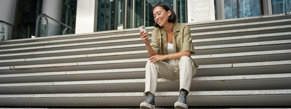 Portrait of happy smiling asian woman, sitting outdoors near building, using smartphone. Technology concept.