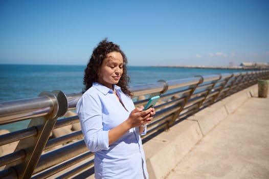 Beautiful multi-ethnic woman in blue casual shirt checking mobile app on her smartphone, smiling, enjoying beautiful sunny day, standing at seascape background.