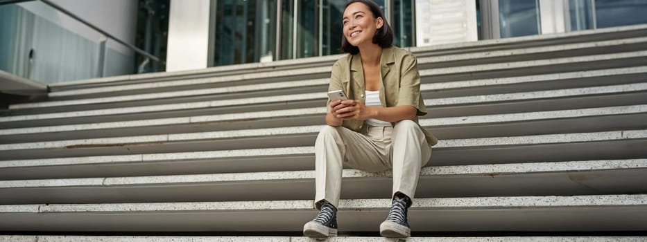 Portrait of happy smiling asian woman, sitting outdoors near building, using smartphone. Technology concept.