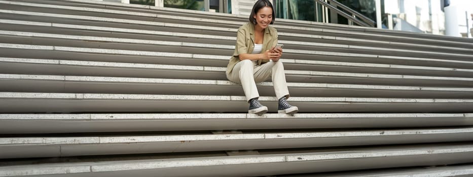 Portrait of smiling asian girl sits on stairs outdoors, sending message, using smartphone app, looking at mobile phone screen.