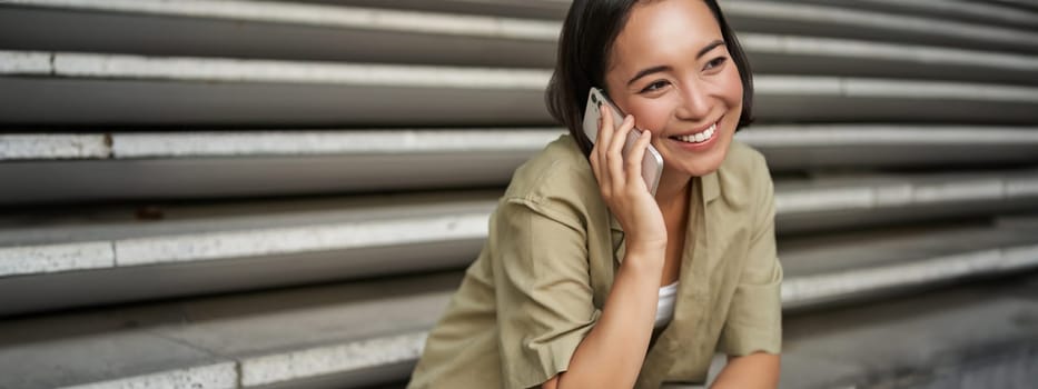 Close up portrait of smiling asian girl talks on mobile phone, sits outside on street stairs. Young woman calling friend on smartphone.