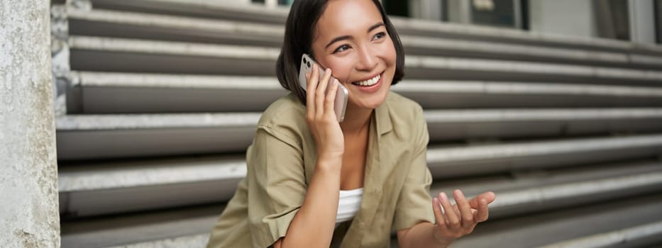 Close up portrait of smiling asian girl talks on mobile phone, sits outside on street stairs. Young woman calling friend on smartphone.
