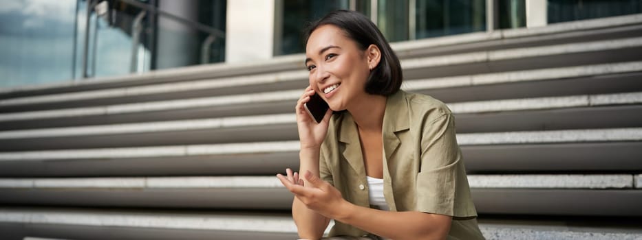 Portrait of beautiful asian girl talks on mobile phone, sits on street stairs. Woman with smartphone smiling, making a call.