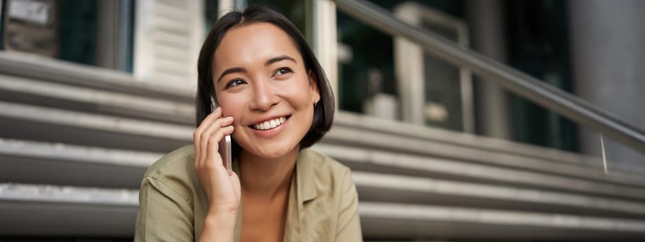 Portrait of asian girl smiles while talks on mobile phone. Young woman calling a friend, sitting on stairs. Technology concept