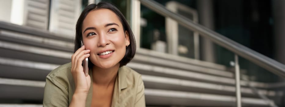 Portrait of asian girl smiles while talks on mobile phone. Young woman calling a friend, sitting on stairs. Technology concept