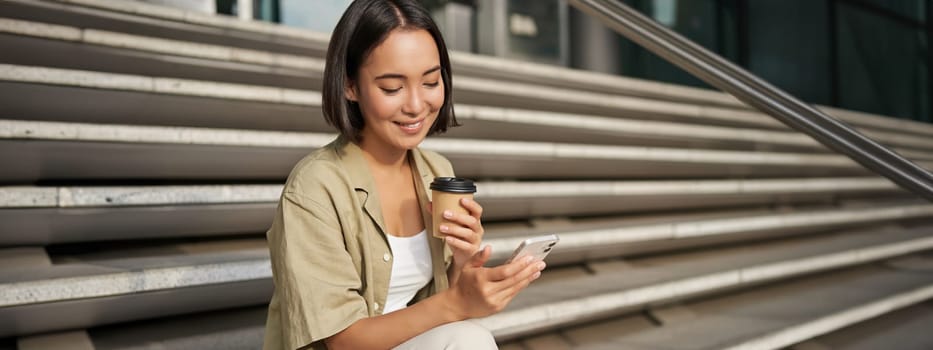 Happy urban girl drinks her takeaway coffee and scrolls feed on smartphone. Asian woman sits on stairs with tea and holding mobile phone.