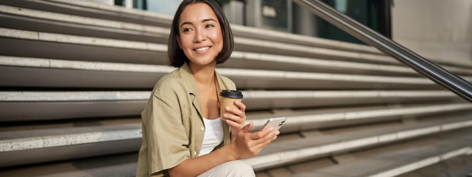 Happy urban girl drinks her takeaway coffee and scrolls feed on smartphone. Asian woman sits on stairs with tea and holding mobile phone.