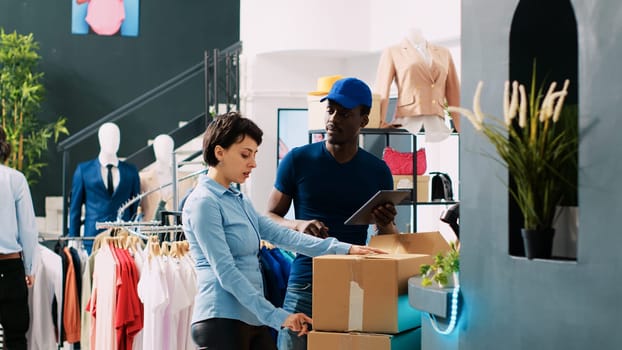 Worker looking at packages with courier, discussing shipping details in modern boutique. African american deliveryman explaining logistics report to store employee in shopping centre.